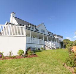A white house with a large porch and railing overlooking a grass lawn with flower beds at Haul A Gwynt in Criccieth