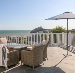 A patio with wicker chairs a glass table and an umbrella overlooking the sea and a distant castle at Haul A Gwynt in Criccieth