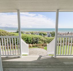 A porch with white railings overlooking a garden with hedges and a view of the sea at Haul A Gwynt in Criccieth