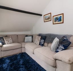 A sitting room with a beige corner sofa with cushions a dark blue rug and framed pictures on the wall at Horseshoe Cottage in Hartland