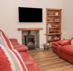A living room with red leather sofas facing a wall-mounted TV and a fireplace with a wooden mantel and a wooden shelf unit at Oak Cottage in Windermere