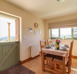 A dining area with a wooden table set for four near a half open green door and a window with cloth blinds at Barcud Cottage in Golden Grove near Llandeilo