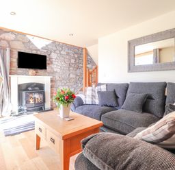 A living room with a gray sofa and pillows a wooden coffee table with flowers a fireplace with a mounted tv and a stone wall at Gwennol Cottage Golden Grove near Llandeilo