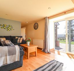 A living room with a dark sofa, wooden coffee table with flowers, wall clock, and sliding glass door at Gwennol Cottage in Golden Grove near Llandeilo