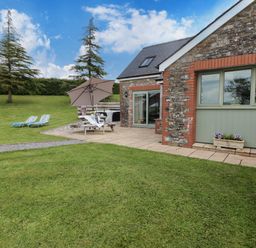 A stone cottage with a patio table and umbrella on a lawn with two lounge chairs and trees at Gwennol Cottage in Golden Grove near Llandeilo