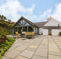 An outdoor patio area with wooden chairs and table next to a house with a glass extension and white walls at Balog Farm in Amlwch