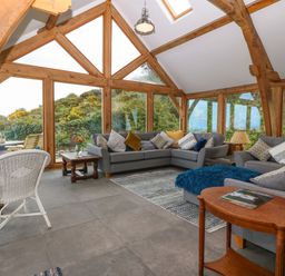 A sitting room with gray sofas pillows a white wicker chair wooden tables and large windows at Balog Farm in Amlwch