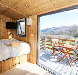 A small wooden bedroom with a bed towels and a wall-mounted television next to an open sliding door leading to an outdoor deck with a table and chairs at Elan in Rhayader