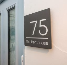 A gray door next to a black sign with white text showing number 75 and The Penthouse