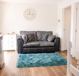 A living room with a sofa and two patterned cushions a blue rug a white clock on the wall and kitchen appliances at Oakleigh in Four Crosses