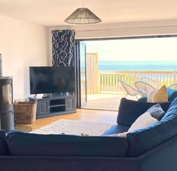 A living room with a blue sectional sofa a flat screen tv on a stand a wood stove and an open door leading to a balcony with chairs and a sea view at Ty Ni in New Quay