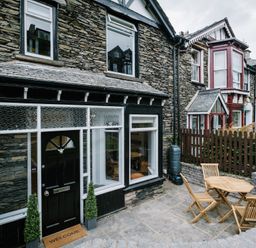 A stone house exterior with a black door and windows and a small outdoor seating area with wooden table and chairs at Craig Cottage in Bowness-On-Windermere