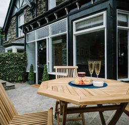 A wooden outdoor table with two glasses of champagne and a plate of scones in front of a stone house at Craig Cottage in Bowness-On-Windermere