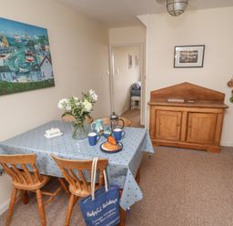 A dining area with a wooden table covered by a blue tablecloth and four wooden chairs a vase with flowers croissants on a plate and mugs on the table a wooden cabinet on the right and a painting on the left wall at 9 Harefield Cottages in Lympstone