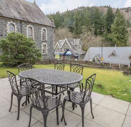 An outdoor patio with a metal table and six chairs overlooking a garden and stone buildings with trees in the background at Ty Capel in Betws-Y-Coed