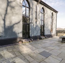 An outdoor stone patio with a table and chairs adjacent to a building with tall arched windows at Greystones Chapel in Cwm-y-Glo near Llanrug
