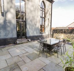 A patio area with a glass table and six metal chairs outside a building with large windows at Greystones Chapel in Cwm-y-Glo near Llanrug