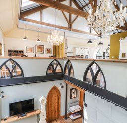 A two-story room with exposed wooden beams chandeliers a TV and a clock at Greystones Chapel in Cwm-y-Glo near Llanrug