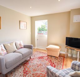 A living room with a gray sofa, a patterned armchair, a beige ottoman, a television on a glass stand, and wall art at Roundham Cottage in West Bay