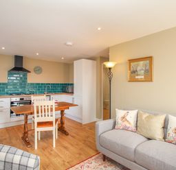 An open plan living room and kitchen with a wooden dining table and chairs and a gray sofa with animal print cushions at Roundham Cottage in West Bay