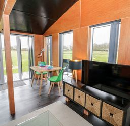 A dining table with green chairs and tableware beside a TV on a black stand with baskets in a room with large windows at The Tractor Shed in Lower Broadheath Worcester