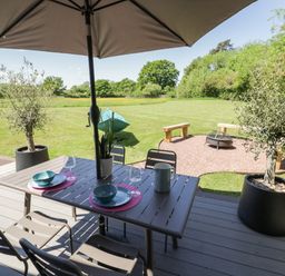 An outdoor dining table with chairs and an umbrella on a deck overlooking a lawn with a fire pit and wooden benches at The Tractor Shed in Lower Broadheath Worcester