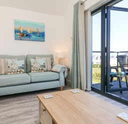 A living room with a light blue sofa and cushions a wooden coffee table and a glass door leading to a balcony with chairs at Seaquel in Westward Ho