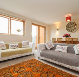 A living room with two sofas cushions a dining table and a large clock on the wall at Maple in Falmouth