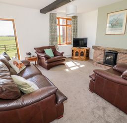 A living room with brown leather sofas a TV on a wooden stand a brick fireplace and windows showing an outdoor field at Manor Farm Cottage Hill Croome near Upton Upon Severn