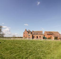 A grassy field with a wooden fence and brick farm buildings under a blue sky at Manor Farm Cottage in Hill Croome near Upton Upon Severn