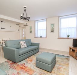 A living room with a blue sofa and ottoman a wooden TV stand with a TV two windows and a dining table with chairs at Bliss On The Bay in Weymouth