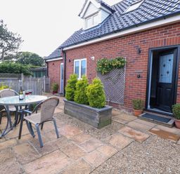 An outdoor patio area with a round glass table, two chairs, a wooden planter with plants, and a brick house exterior at Spinney's Den in Knapton