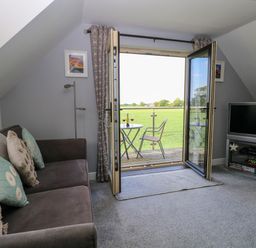 A living room with a sofa pillows and a TV near open double doors leading to a balcony with a table and chairs overlooking a field at Spinneys Den in Knapton