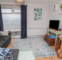 A living room with a sofa and cushions a television on a table and a large window with curtains showing a sea view at Sea Snug in Spittal