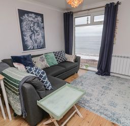 A living room with a dark sofa with patterned cushions a small table a rug and a large window with a sea view at Sea Snug in Spittal