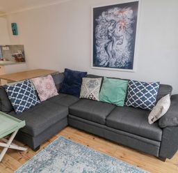 A gray sectional sofa with patterned cushions in a living room with wooden floor and a kitchen in the background at Sea Snug in Spittal