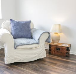 An armchair with a blue cushion and throw next to a wooden box with a lamp on it in a room at 22 Gwel Yr Wyddfa in Menai Bridge