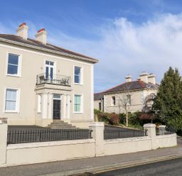 A two-story house with a balcony and black front door surrounded by a low wall and trees at Beachcomber in Ballycastle
