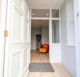 An open white front door leading to a hallway with wooden flooring a red chair with a yellow footstool and a floor lamp Beachcomber Ballycastle