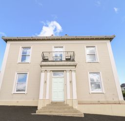 A two-story beige building with six windows and a white front door with columns and a small balcony above at Beachcomber in Ballycastle