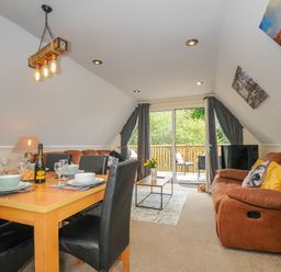A dining table with place settings and a bottle next to a living area with brown sofas and a sliding door to a balcony at Dart Lodge in Callington