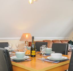 A dining table set with bowls plates glasses cutlery a bottle of prosecco and croissants with chairs sofa and lamp in a room at Dart Lodge in Callington