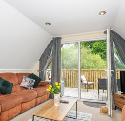 A living room with brown sofas a coffee table with yellow flowers and a TV by a sliding door leading to a balcony with chairs at Dart Lodge in Callington