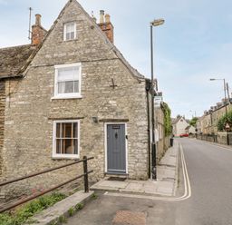 A stone cottage with a gray door and two windows on a corner next to a street with more stone houses at Corner Cottage in Malmesbury