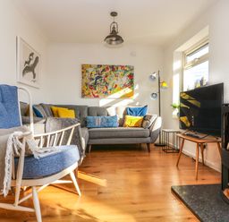 A living room with a blue armchair cushions on a gray sofa a flat screen TV and a wood burning stove at Rock Pool House in Porthleven