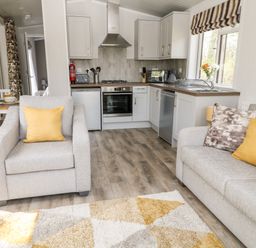 A living area with armchair and sofa with yellow cushions and a kitchen with white cabinets at Dove Lodge Highampton near Sheepwash