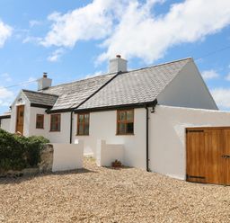 A white cottage with a gray tiled roof and wooden doors beside a gravel driveway at Plas Coch Rhoscolyn in Rhoscolyn