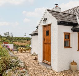The exterior of a white cottage with a wooden door and windows surrounded by gravel garden with plants and outdoor seating at Plas Coch Rhoscolyn