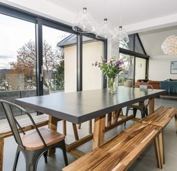 A dining area with a table, benches, metal chairs, and a vase with flowers next to large glass windows at Kessock in Bowness-On-Windermere
