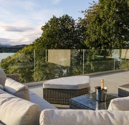 An outdoor seating area with a cushioned sofa, ottoman, glass table with a bottle in an ice bucket and glass on a terrace with glass railing overlooking trees and water at Kessock in Bowness-On-Windermere
